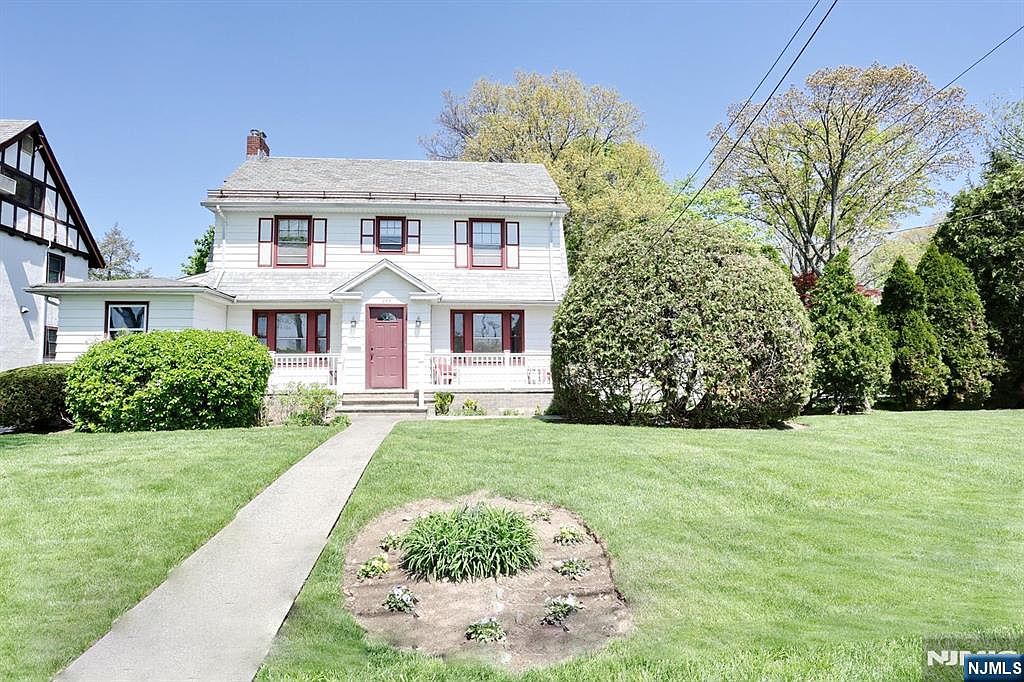 Front elevation of a white clapboard center-hall Colonial with deep-red front door, red shutters, slate-grey roof and a covered front porch, set on a manicured spring lawn under a blue sky